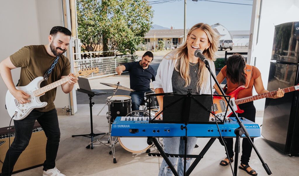Female singer playing keyboard and performing with a guitarist, bassist, and drummer in a bright studio.